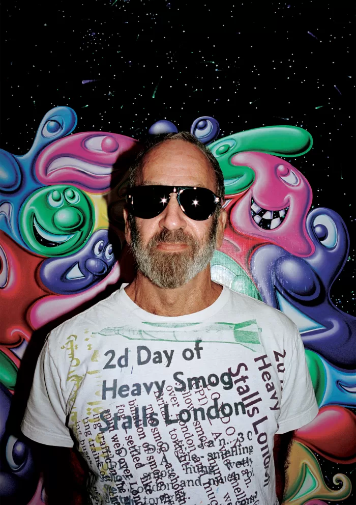 A photograph of Kenny Scharf stood in front of one of his paintings