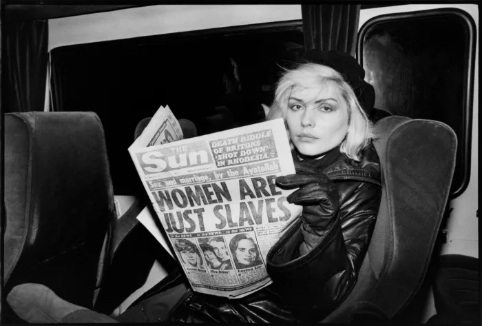 A black and white photograph of Debby Harry holding The Sun newspaper