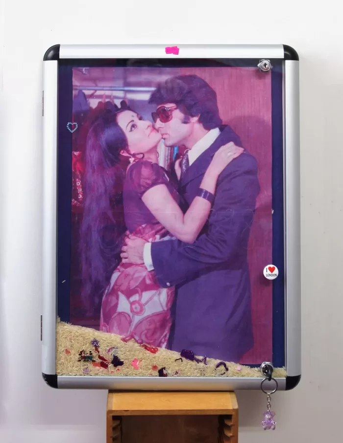 A photograph of a kitsch Y2K style moodboard of a picture of a couple embracing overlayed with stickers and trinkets. The moodboard is framed and the bottom of it is filled with rice