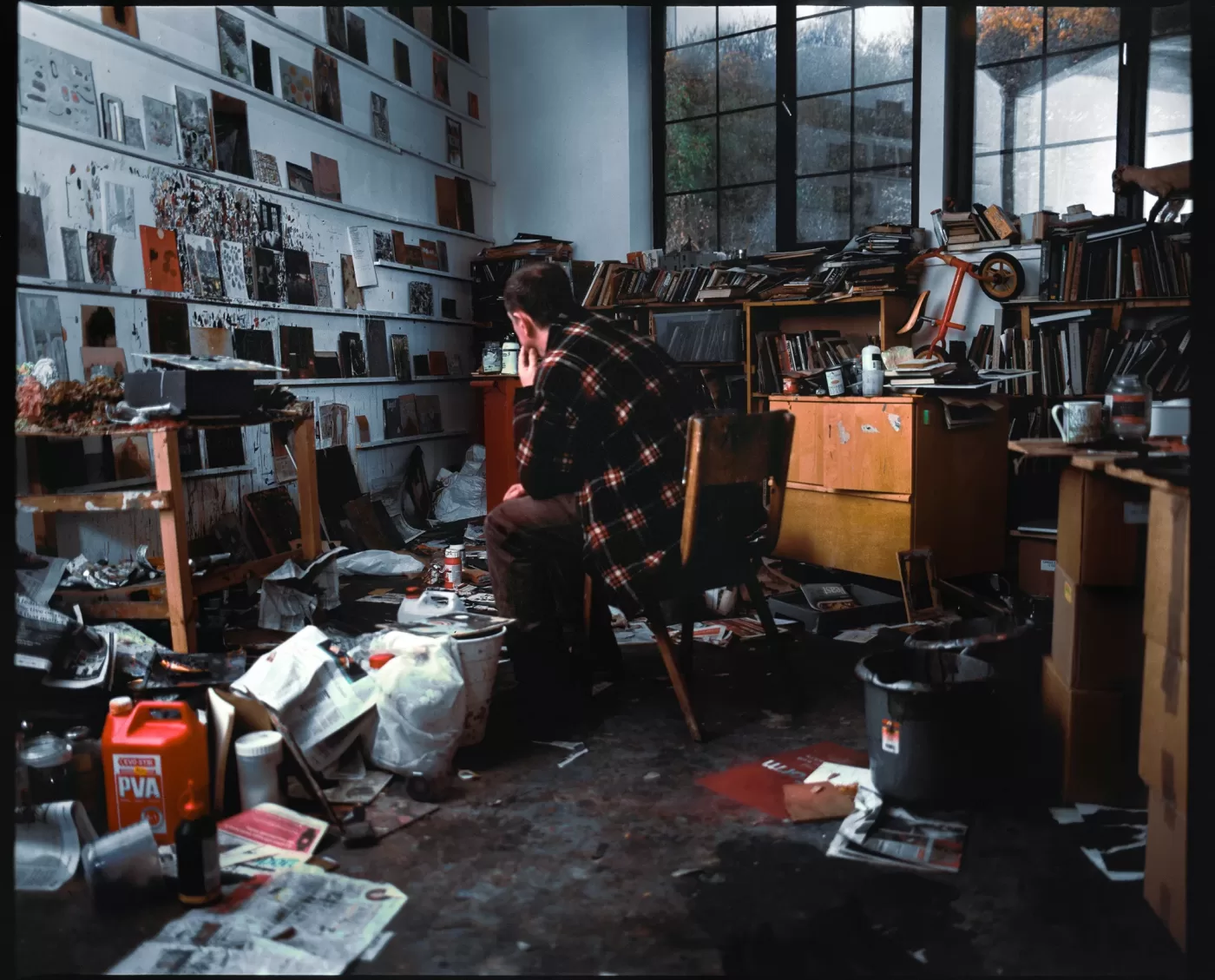 A photograph of Andrew Cranston sat at his desk in his studio. The room is full of artwork on shelves and on the floor