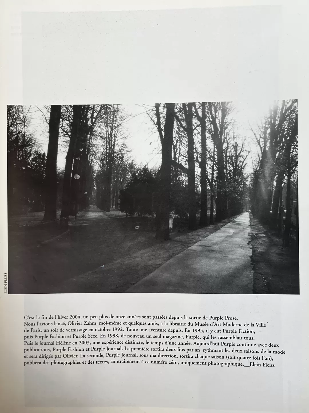 A photograph of the page of a photobook picturing a black and white photograph of a road surrounded by trees