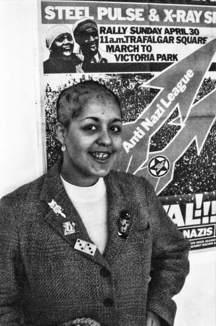 Black and white photograph of a woman smiling, stood in front of an Anti Nazi League poster