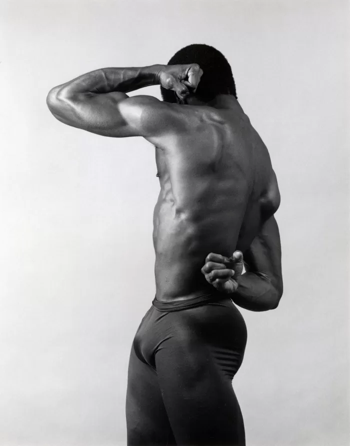 A black and white photograph of Alistair Butler taken by Robert Mapplethorpe. He is posed topless flexing his muscles