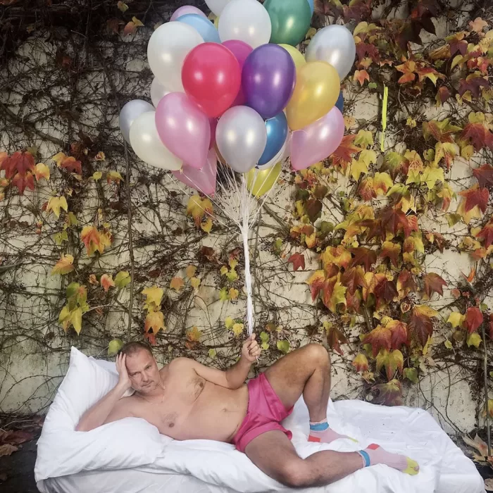 A photograph of Juergen Teller in underwear on a mattress holding a bunch of balloons