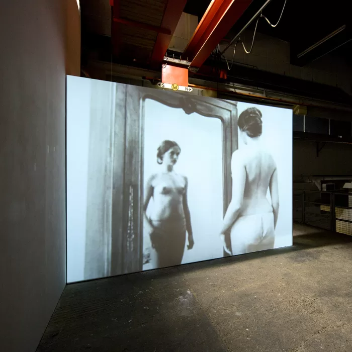 Installation shot of a Chantal Akerman film at Ambika P3 London which shows a half naked female standing in front of a mirror.