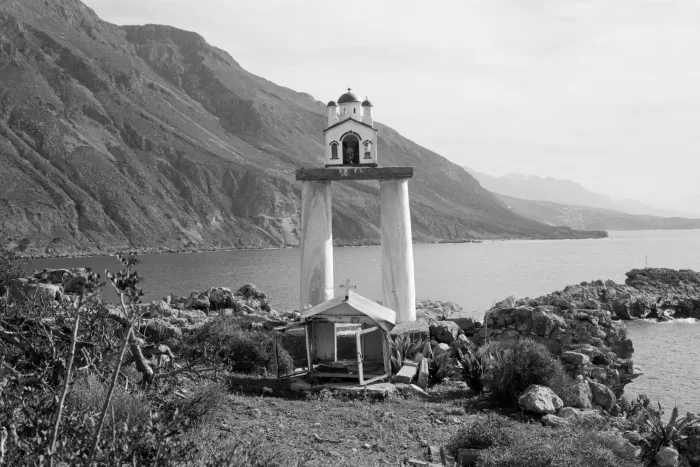 a black and white photograph taken by Daniel Adhami of an devotional and commemorative icon station taken in Crete, Greece overlooking a mountainous sea-scape