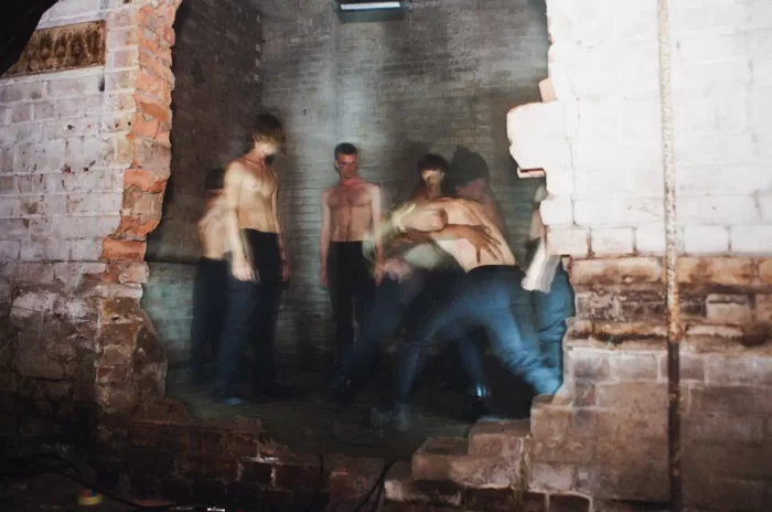 A blurry flash photograph of the performers as they stand shirtless looking towards eachother